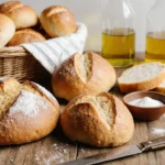 A rustic wooden board holding a freshly baked artisan loaf, a perfect sandwich bread loaf, and warm dinner rolls, with a bread knife and flour sack cloth.