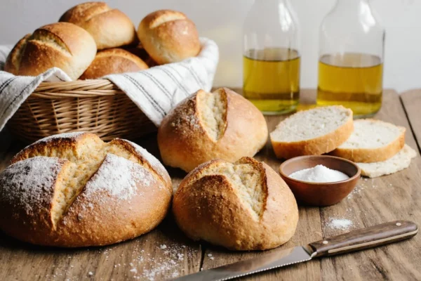 A rustic wooden board holding a freshly baked artisan loaf, a perfect sandwich bread loaf, and warm dinner rolls, with a bread knife and flour sack cloth.